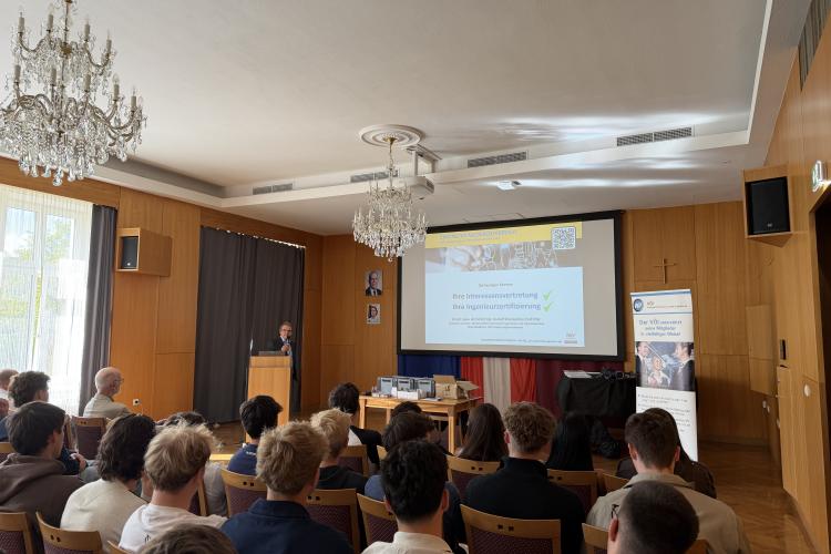 Vortragender hält Präsentation über Interessensvertretung und Ingenieurzufriedenheit vor Publikum in einem Saal mit Kronleuchtern.